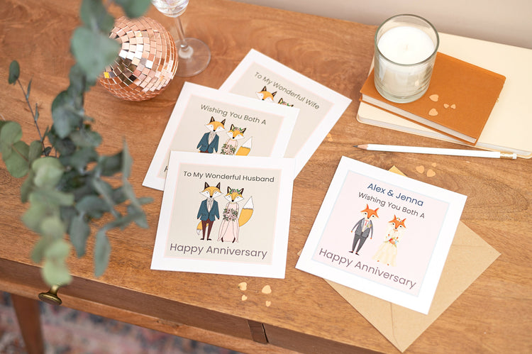 A group picture of a selection of wedding anniversary cards, ones for wife's, husbands and couples. The cards feature cartoon style mr and mrs fox characters. The cards are on a table with stationery and green foliage.