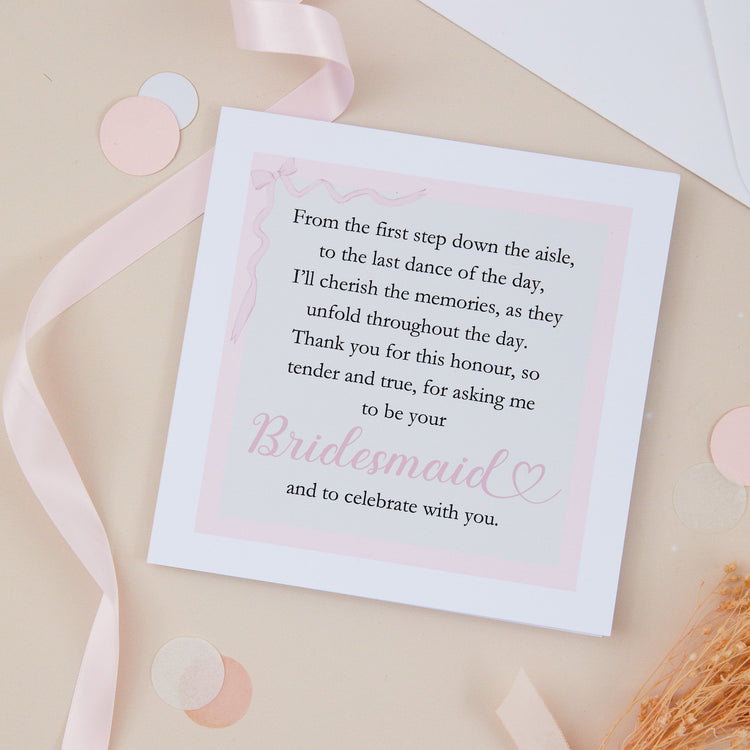 A thank you for asking me to be your bridesmaid card. Set in the form of a poem, against a pale background and pink border. The card is set against a pale pink background with stationery, and envelope and dried flowers.
