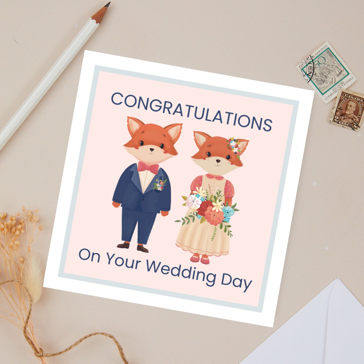 Wedding Congratulations card with a Baby fox couple, he is wearing a blue suit with a red bow tie, and she is Wearing a beige dress without sleeves, and holding a floral bouquet. She also has a small floral arrangement in her hair. They are set against a pink background with a blue border. There is stationery in the background against a beige background.