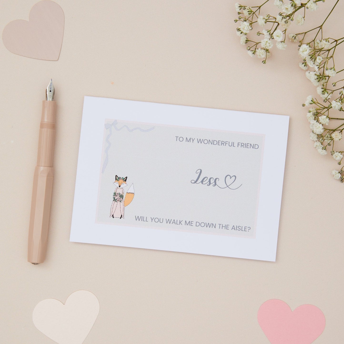 To My Wonderful Friend will you walk me down the Aisle proposal Card. A Girl fox in wedding bridal attire on a soft pink background with a ribbon border, with the label 'To my wonderful friend, will you walk me down the aisle. The Card is set against a pale beige background with stationery items.