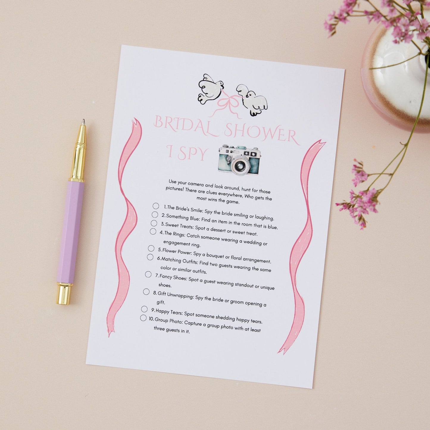 A 'I Spy' game card for bridal shower or hen do parties, with pink accents and a Camera. There is a pen for writing, displayed on a table with a floral decoration.