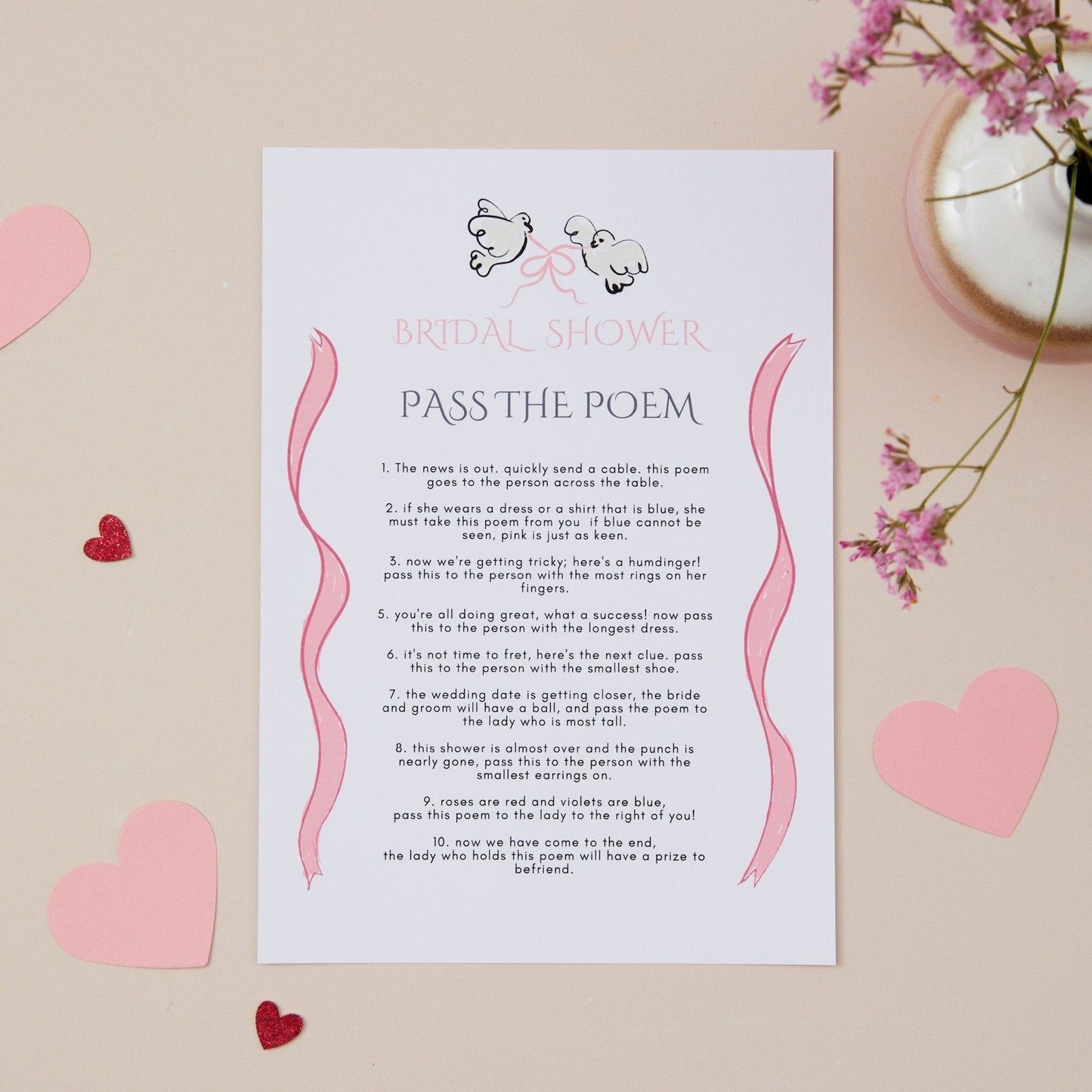 A bridal shower game card titled 'PASS THE POEM' with a heart and ribbon design, placed on a pink background surrounded by heart-shaped decorations and floral elements.