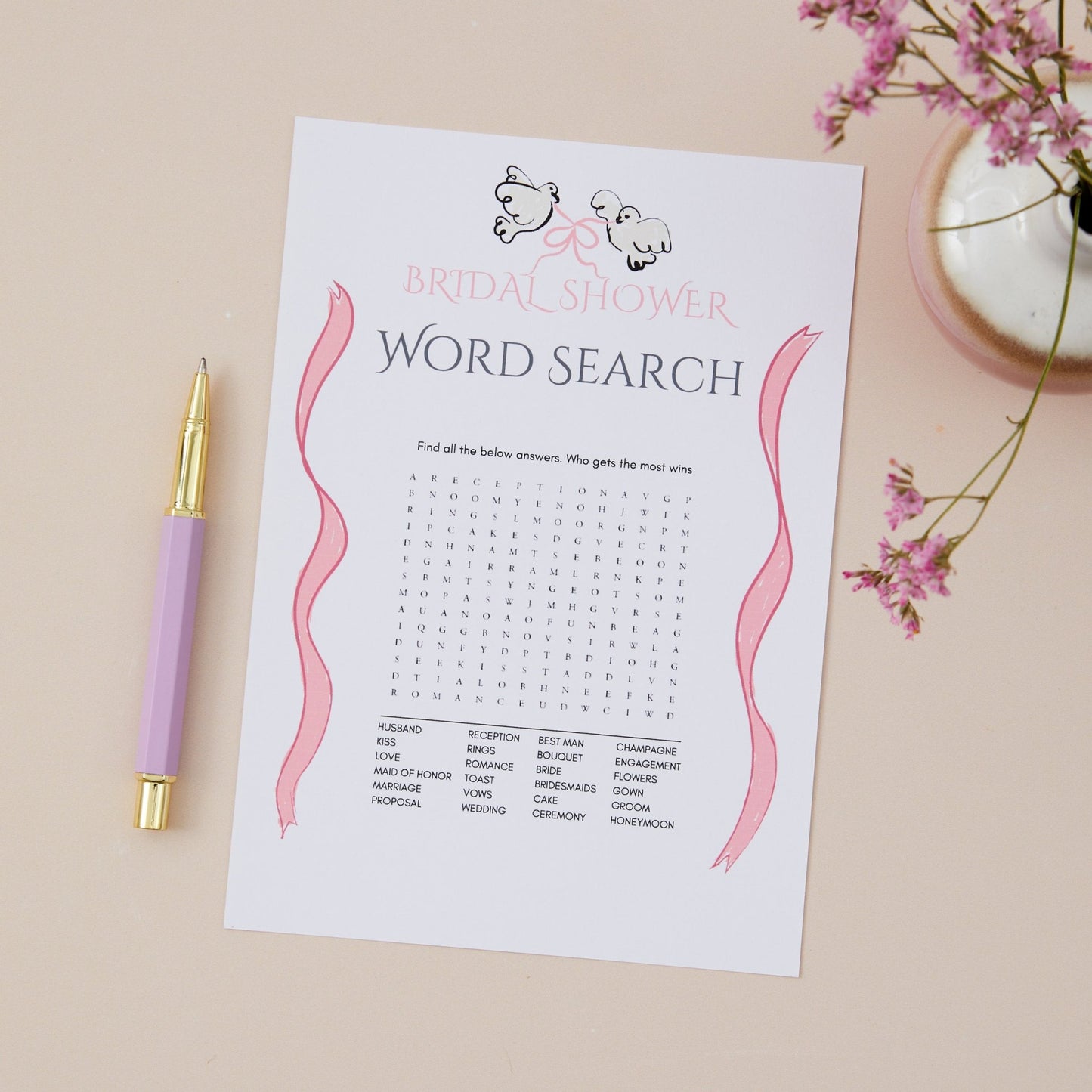 A printed 'Bridal Shower Word Search' game sheet showing a word search grid and words to find at the bottom of the page card. the background has a pen and decorative flowers on a table.