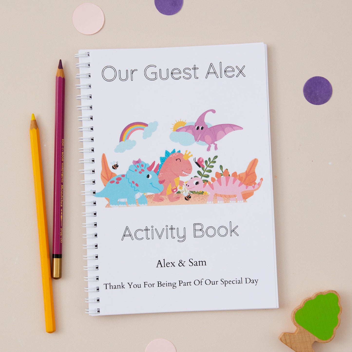Dinosaur Characters Children’s Wedding Activity Book Pack. An A5-sized children's wedding activity book titled 'OUR GUEST ALEX' with a colorful dinosaur illustration on the cover, placed next to a pencil and some decorative stickers.