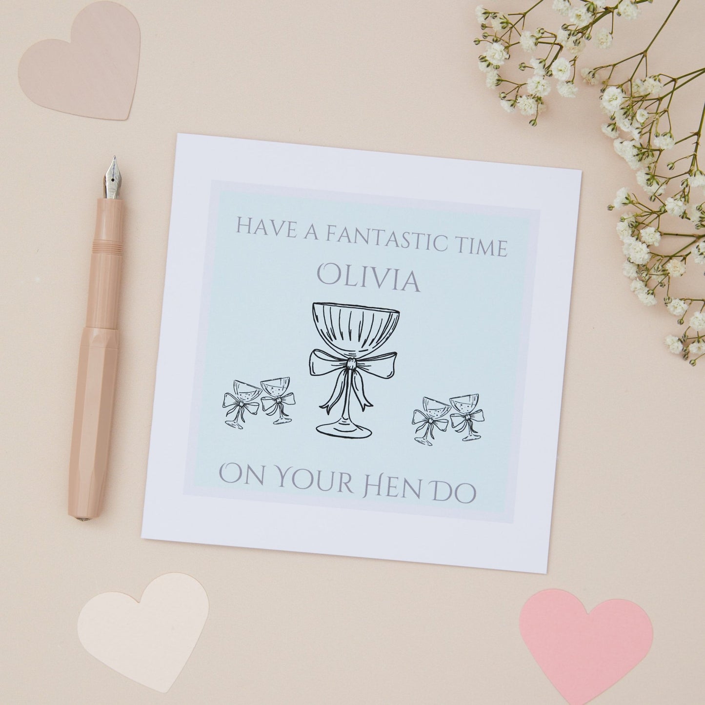 A hen do bridal shower party card with a message 'HAVE A FANTASTIC TIME ON YOUR HEN DO' featuring a drawing of celebratory glasses. The card is displayed on a neutral background with some decorative elements like a pen, heart-shaped cutouts, and floral branches.