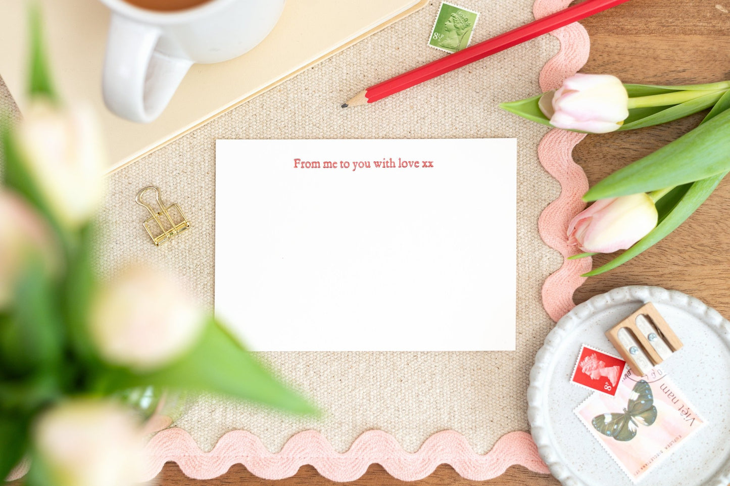 A flat letterpress notelet card with the message 'From me to you with love' printed on it, displayed on a desk with a cup of tea, pens, and tulips.