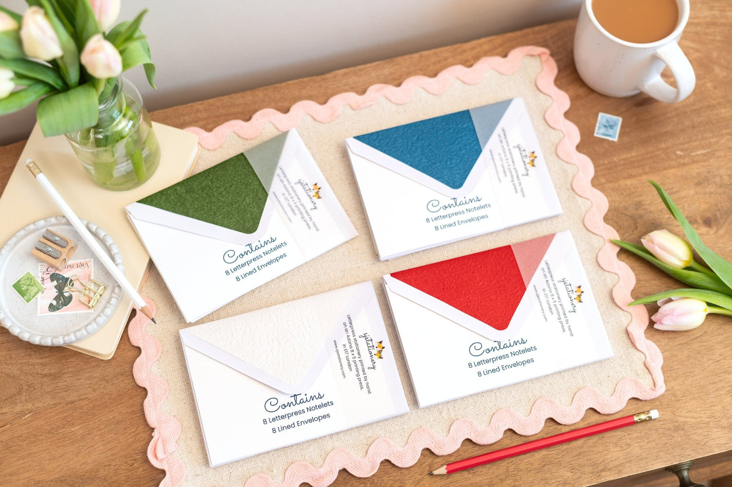 Envelopes which are green,blue.red, white shown as colour options to accompany letterpress cards. The coloured envelopes are placed on a scalloped table cloth with stationery and a cup of tea, plus tulips.