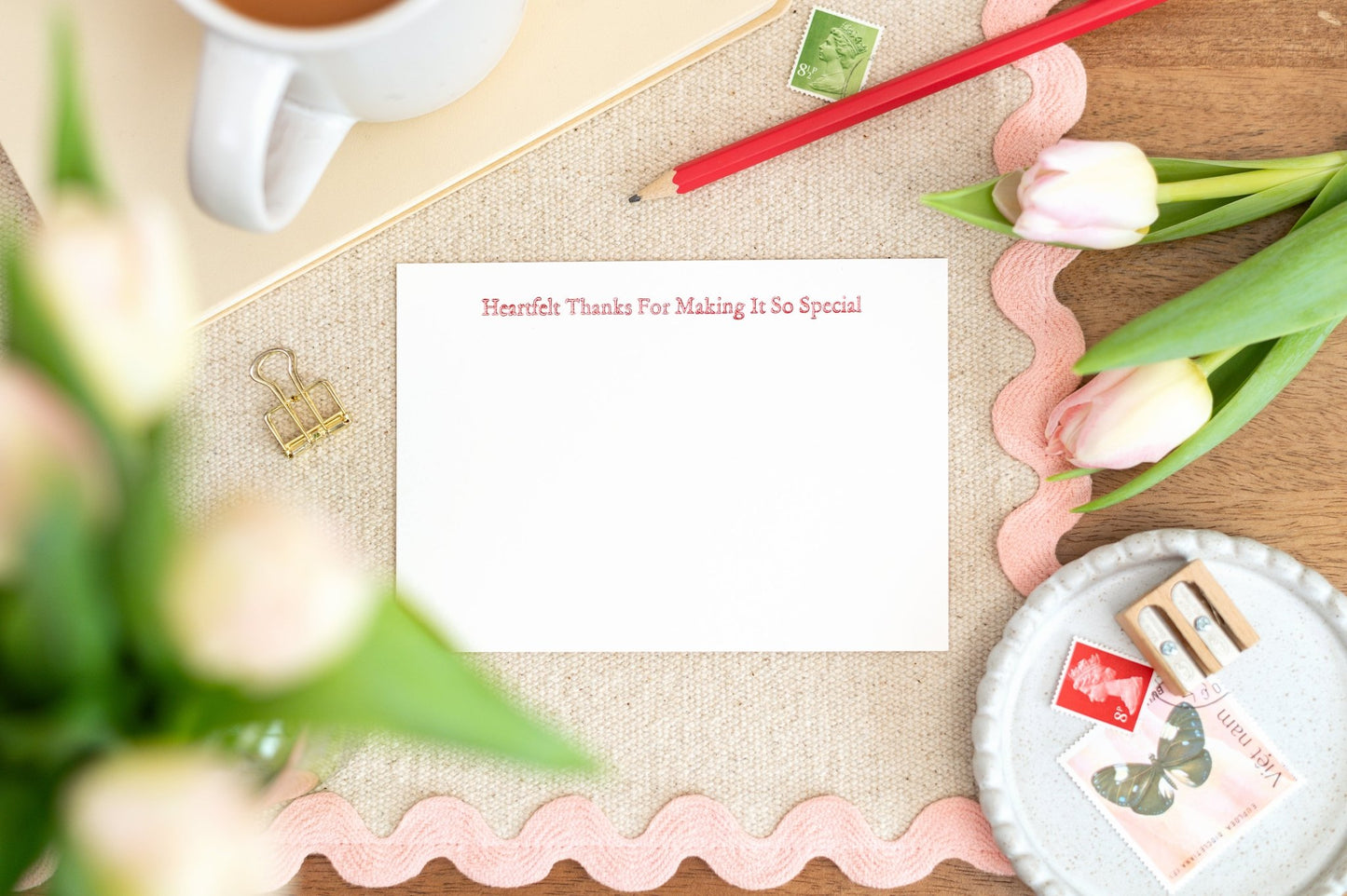 A flat lay image of a letterpress thank you notelet card set with a message that reads 'Heartfelt Thanks for Making It So Special'. The set is displayed with tulips, a cup of tea, and writing utensils.