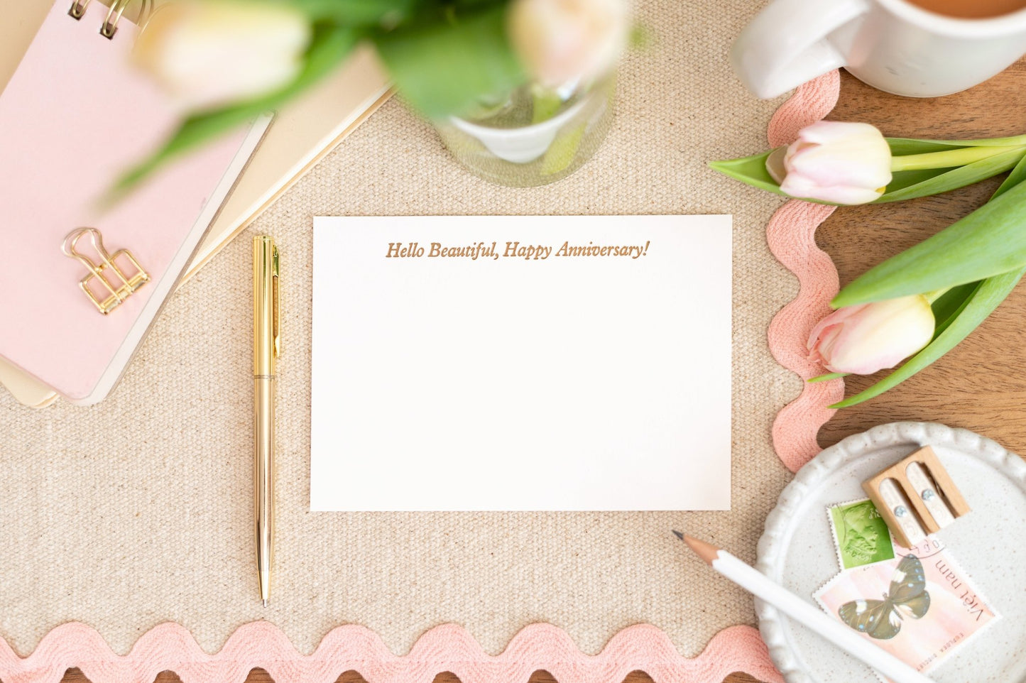 Letterpress Printed Notelet Card with the words 'Hello Beautiful, Happy Anniversary!' in gold writing. Is set against a decorative table setting with flowers and stationery