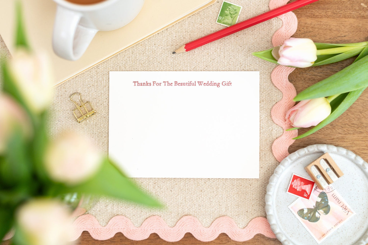 A flat letterpress notelet with the message 'Thanks for the beautiful wedding gift' printed in classic letterpress typography, displayed on a desk with a cup of tea, pens, and tulips.