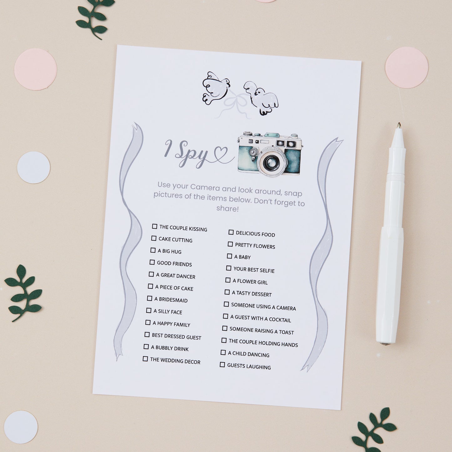A 'I Spy' wedding reception game card with written suggestions for items to find at a wedding, placed on a table with decorative elements and a pen.
