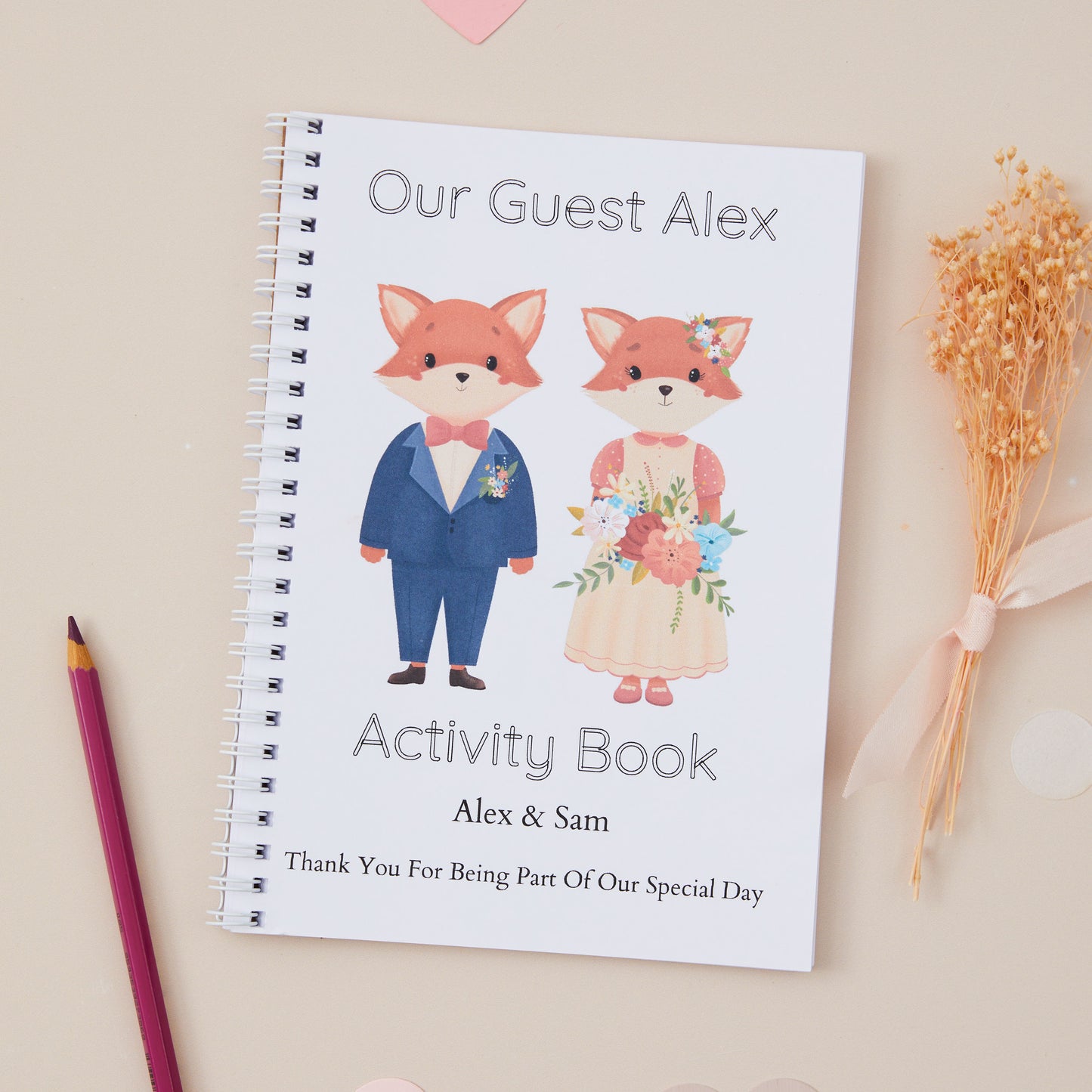 Kids Wedding Activity Pack Book - featuring a fox baby couple dressed in wedding attire as the themed illustration on the front with the title 'our guest alex activity book'. There is also the names of the couple and the words 'thanks for being part of our special day'. The Activity book is set on a beige background with dried flowers and stationery items.