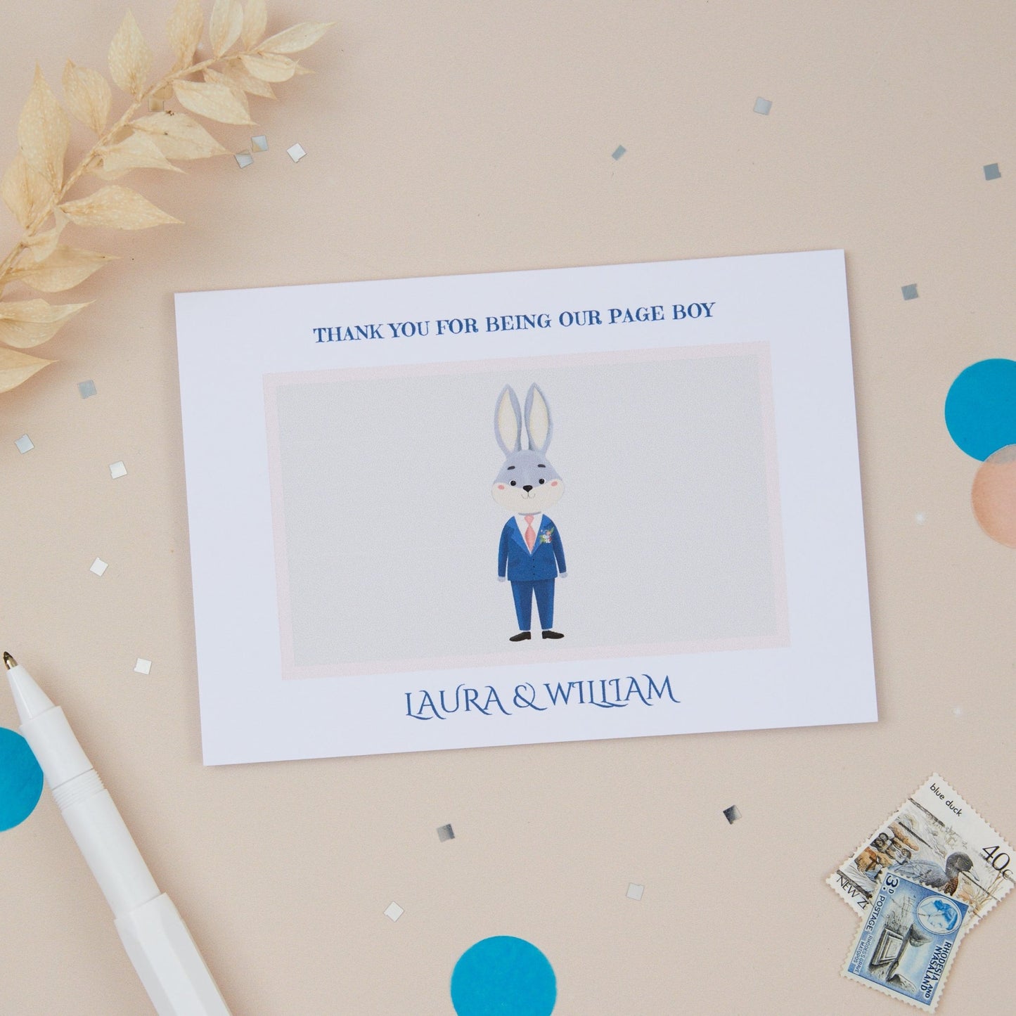 A 'Thanks for being our Page Boy' card with a illustration of a rabbit in a blue wedding suit, placed on a neutral background with some decorative elements like blue confetti, dried flowers and stationery items.