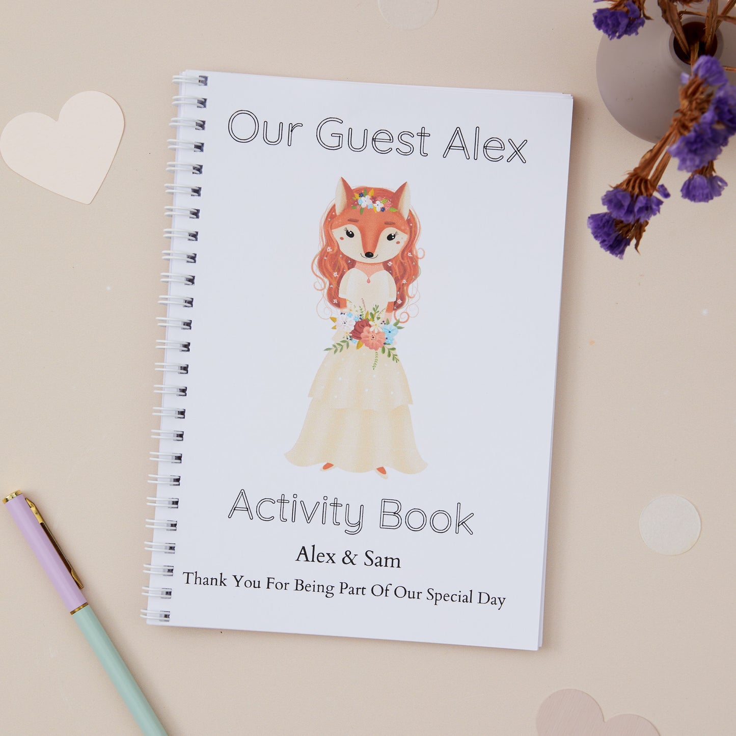 Children's Wedding activity book with an Auburn haired girl fox dressed in bridal dress. She is holding a floral bouquet.
The illustration is on a beige background with stationery and purple dried flowers in the background