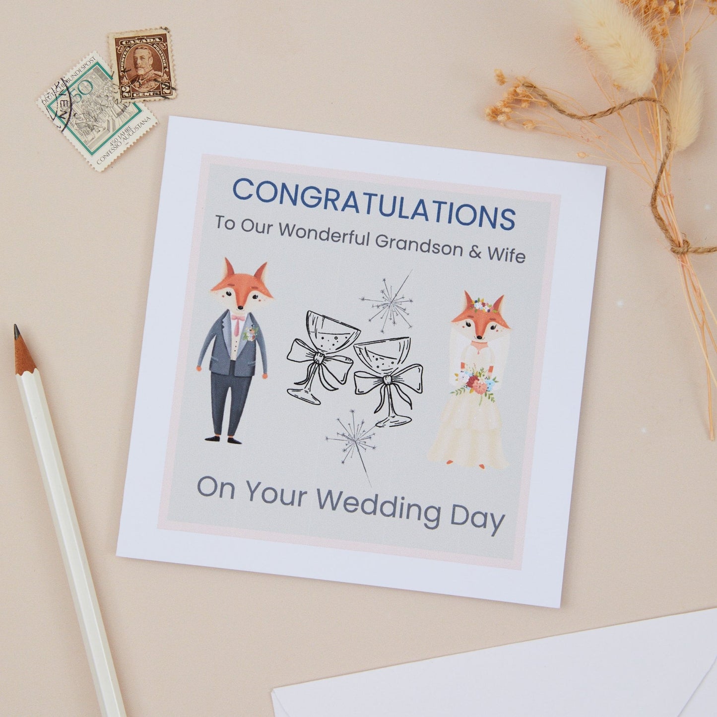 A greeting card with a cartoon illustration of a Fox bridal couple and text that reads 'Congratulations To Our Wonderful Grandson & Wife On Your Wedding Day'. The Bridal couple are separated by two celebratory champagne classes with bows. The card is displayed on a neutral background with a pen and vintage stamps.
