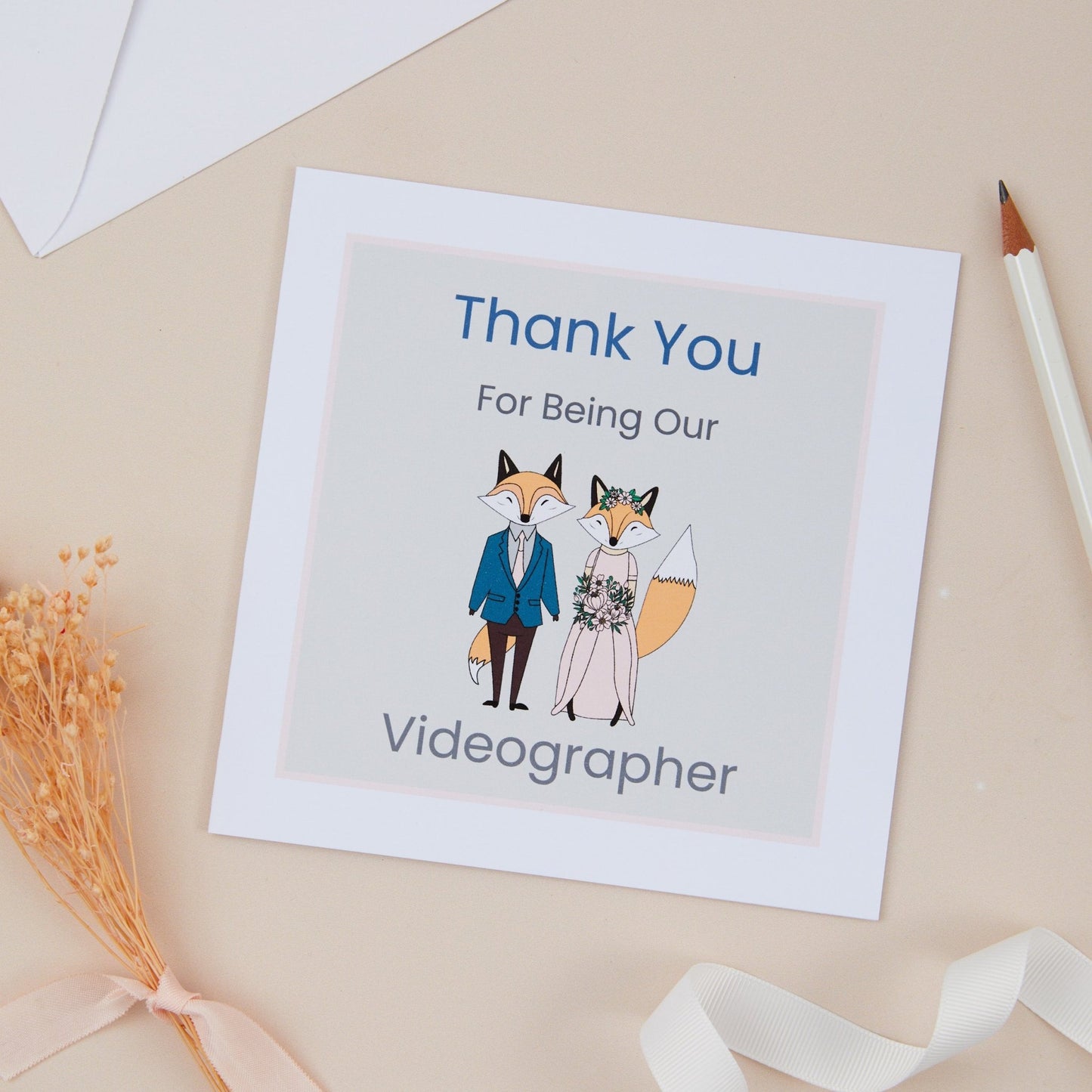 A 'Thank You for Being Our Videographer' card with an illustration of a fox couple. The foxes are placed on a neutral background with a pink border. The Card is set against a beige background with a pen, ribbon, and dried flowers.