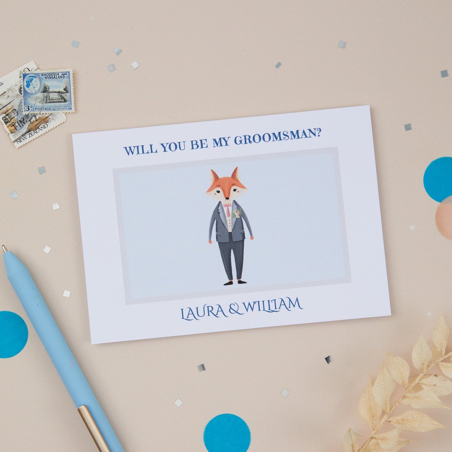 A6 sized card with a cartoon illustration of a fox in a suit, with the text 'WILL YOU BE MY GROOMSMAN?' above a white space. The card is displayed with blue confetti, a pen, and a lined envelope.