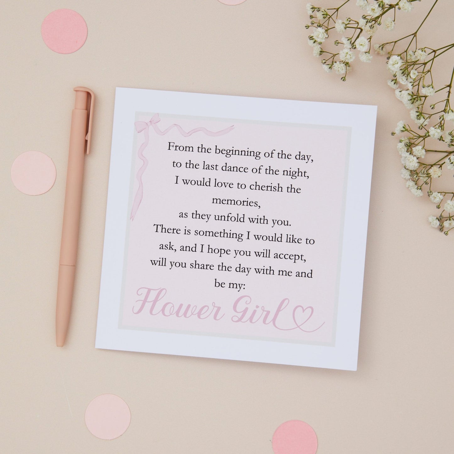 A white card with pink text that reads 'Will you be my Flower Girl?', text is in the form of a poem. The card is accompanied by a pink envelope, a pen, and some pink decorative elements, displayed on a neutral background.