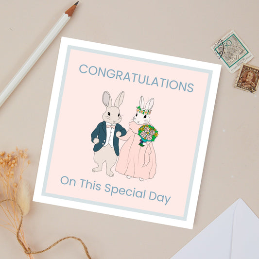 Wedding card congratulations card for a couple with bunnies dressed to get married on a pink card background. The card is surrounded by stationery items. on a beige background.