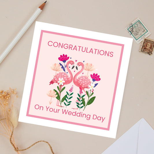 Congratulations on  your Wedding day card with flamingo design - 2 pink flamingos, looking at each other. There are green floral shoots and pink flowers that surround them. The card has a pink background, and a deep pink border. The background is light beige with stationery in the background.