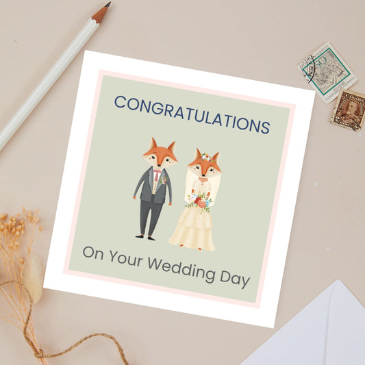 Wedding Day Congratulations card with 2 bridal foxes, a man in grey suit, and woman in her bridal dress holding a floral arrangement. They are set against a light green background on the card. The Text reads - congratulations on your wedding day. The card is set against a beige background with stationery.