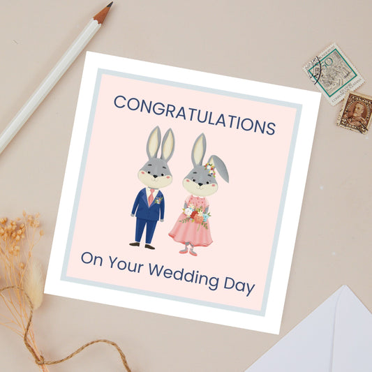Wedding day card with cartoon bunnies, Boy dressed in dark navy suit and girl dressed in light pink dress and is holding a floral arrangement. The card reads 'Congratulations on your wedding day' text on a beige background.