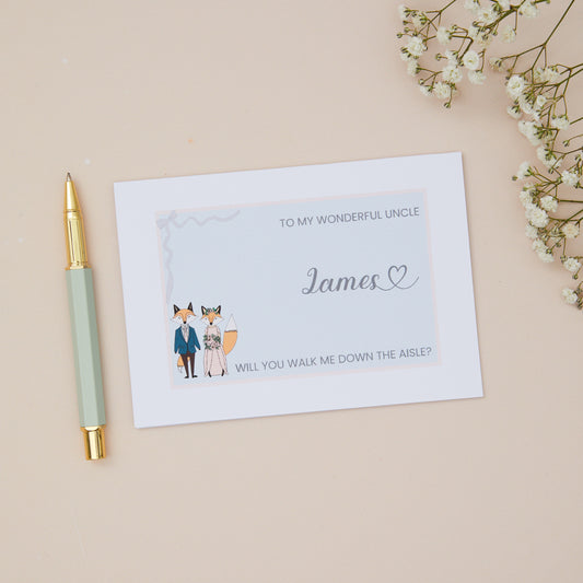 To My Wonderful Uncle will you walk me down the Aisle proposal Card. A  fox couple in wedding bridal attire on a soft pink background with a ribbon border, with the label 'To my wonderful uncle, will you walk me down the aisle. The illustration and text is set against a blue background. The Card is set against a pale beige background with stationery items.