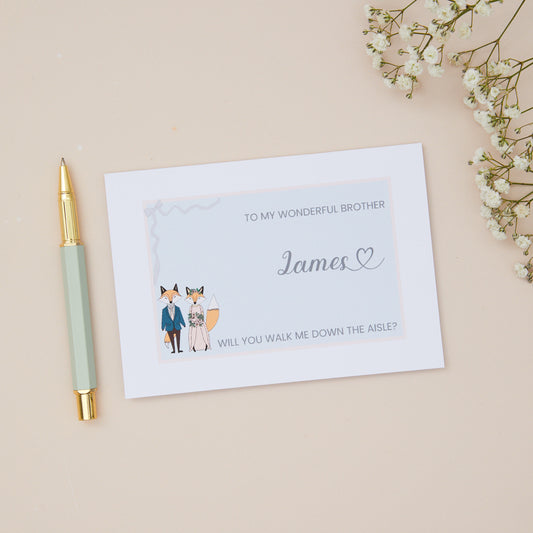 Brother will you walk me down the Aisle proposal Card. A Fox couple with bridal attire with the label 'To my wonderful Brother, will you walk me down the aisle. The Card is set against a pale beige background with stationery items.