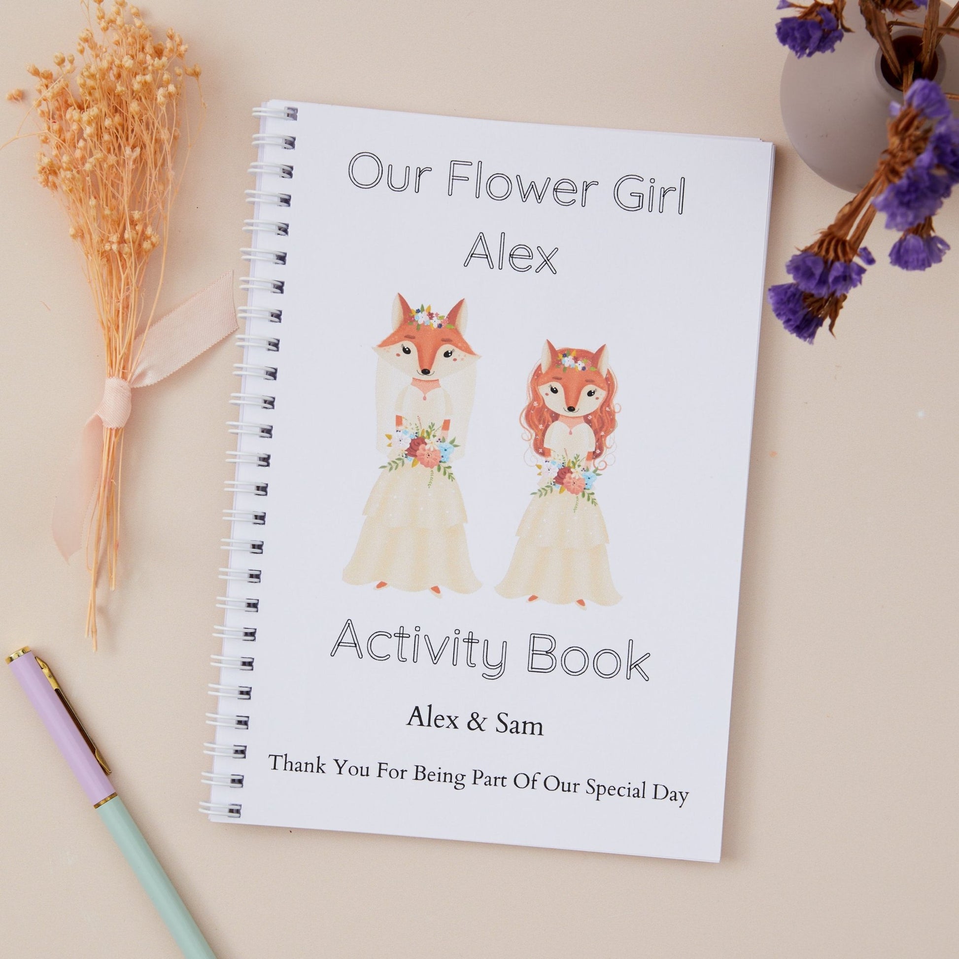 Adorable Fox Bride & Flower Girl Wedding Activity Book Pack. Personalised  cartoon bridal fox and bridal flower girl  on the cover, titled 'Our Flower Girl Alex' with a 'Thank You for being part of our special day' message. The Activity book also has the names of the couple getting married. The activity book is placed on a table with dried flowers and pens.