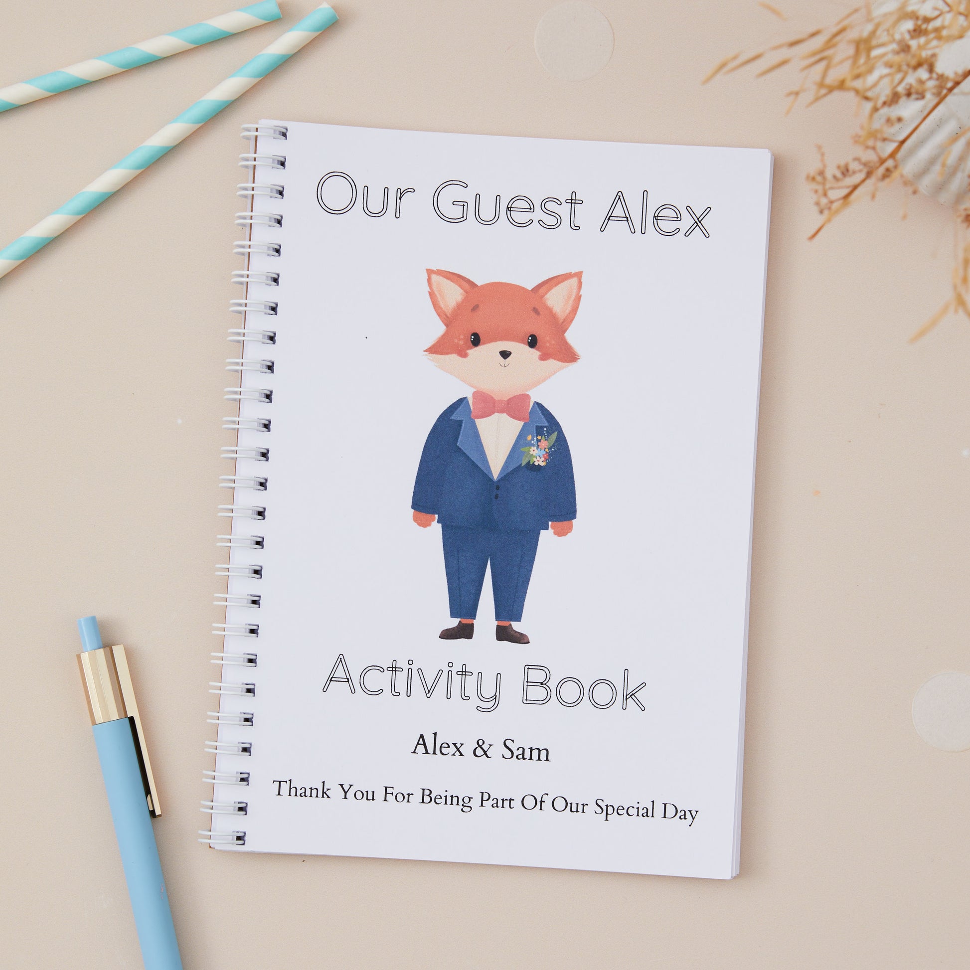 A Blue-Suited Fox Wedding Activity Book Pack For Kids  with a cute fox design, labeled 'Our Guest Alex activity book' with the names of the couple and the label 'thanks for being part of our special day. The activity book is set against a light beige background. There is a Boy Baby Fox in the centre of the book, he is wearing a bow tie and blue suit.