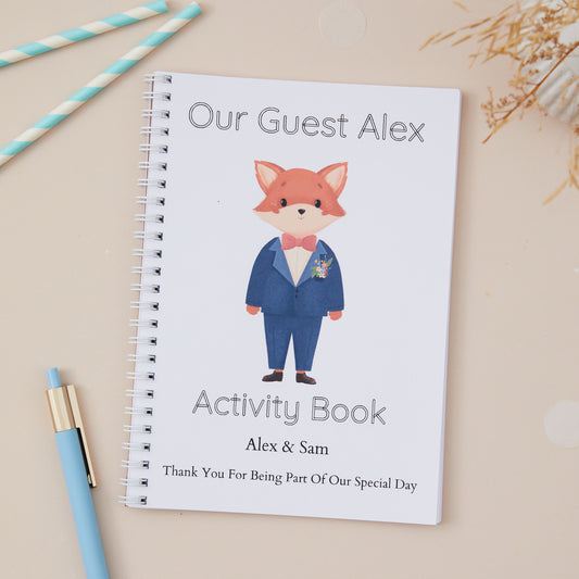 A Blue-Suited Fox Wedding Activity Book Pack For Kids  with a cute fox design, labeled 'Our Guest Alex activity book' with the names of the couple and the label 'thanks for being part of our special day. The activity book is set against a light beige background. There is a Boy Baby Fox in the centre of the book, he is wearing a bow tie and blue suit.