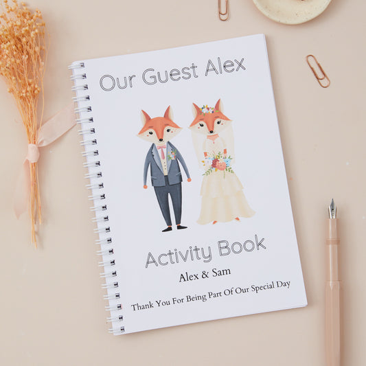 A personalised Bridal Fox Wedding Activity Book.  A5-sized titled 'Our Guest Alex' with a cartoon Bridal fox couple on the cover, alongside a pen and some dried flowers. The name of the couple getting married is also shown, and the label 'thanks for being part of our special day' is underneath the names of the couple. The Wedding Activity Book is set against a light beige background, with stationery items such as paper clips & pens, and there is also some dried flowers.