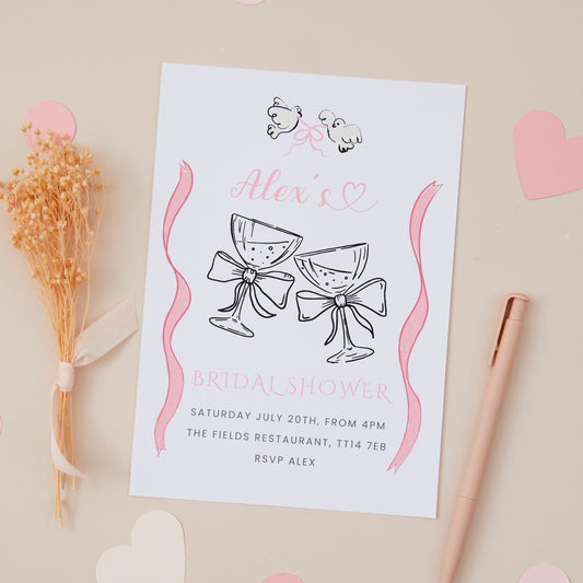 A bridal shower hen do invitation featuring two glasses of champagne with decorative elements in pink and white colors. The invitation is placed on a surface with a pen, decorative dried flowers, and paper hearts around it.