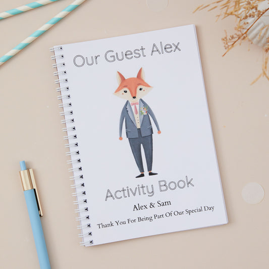 Dapper Grey-Suited Fox Children's Wedding Activity Book Pack 'Our Guest Alex Activity Book' is the text on the front. With a Dapper Grey-Suited Fox character design on the cover, alongside a blue pen and decorative elements. Option to add the name of the couple and their wedding date.