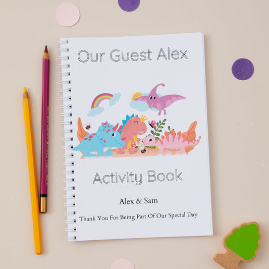 Dinosaur Characters Children’s Wedding Activity Book Pack. An A5-sized children's wedding activity book titled 'OUR GUEST ALEX' with a colorful dinosaur illustration on the cover, placed next to a pencil and some decorative stickers.