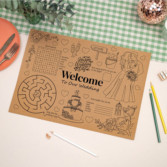 Wedding-themed placemat made with Kraft Brown paper with wedding related colouring - wedding cake, a bride and groom, mazes, word search and puzzles. This placemat is on a checkered tablecloth which has pencils and also a knife and fork.
