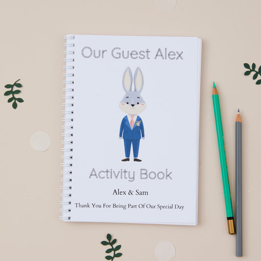 Fluffy Blue Bunny Wedding Activity Book  featuring a boy baby rabbit with floppy ears dressed in wedding attire as the themed illustration on the front with the title 'our guest alex activity book'.  There is also the names of the couple and the words 'thanks for being part of our special day'. The Activity book is set on a beige background with pencils and dried leaves.