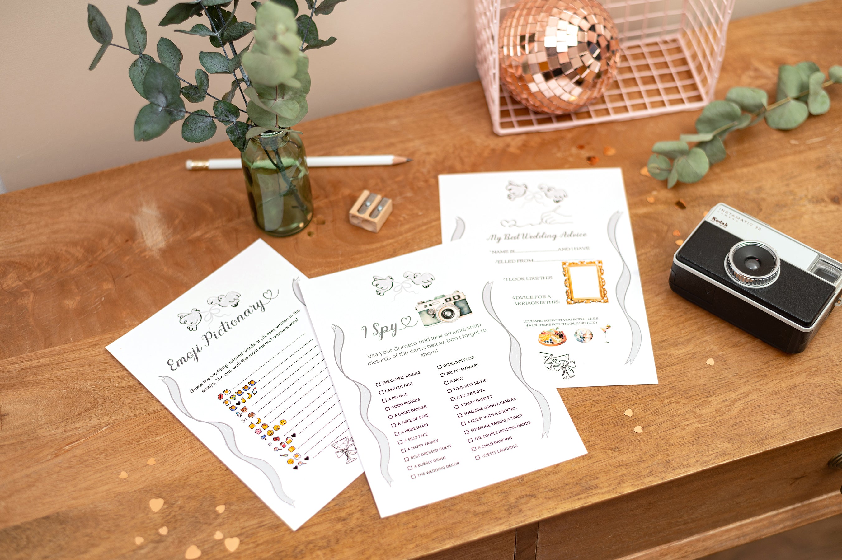 Group photo of 3 different wedding games, I spy, Emoji Pictionary and my best wedding advice. The game cards sit next to a vintage camera on a desk with a green foliage vase, glitter ball and pink decorative wire basket