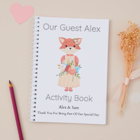 Personalised Little Fox Guest Girl Wedding Activity Book pack for kids  alongside a pen and some dried flowers, on a table with decoration.
