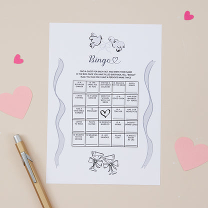 A Bingo game card designed for wedding receptions, with spaces to mark off wedding-related events, accompanied by a pen and decorative heart shapes in the background.