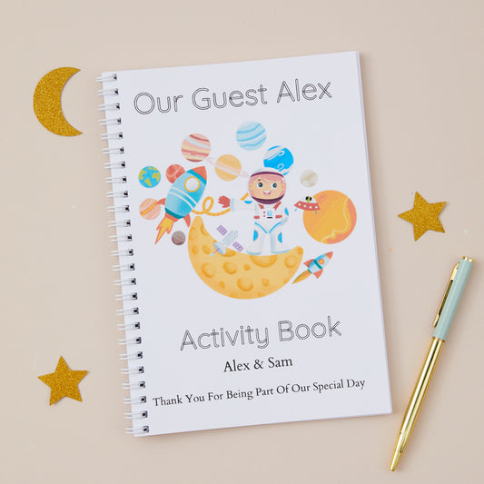 Stargazer Astronaut Wedding Activity Book Pack. Featuring an astronaut themed cover illustration with the title 'our guest alex activity book'.  There is also the names of the couple and the words 'thanks for being part of our special day'. The Activity book is set on a beige background with astrological stars and moon shapes with stationery items.