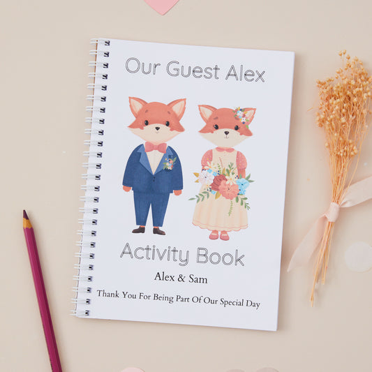 Kids Wedding Activity Pack Book - featuring a fox baby couple dressed in wedding attire as the themed illustration on the front with the title 'our guest alex activity book'.  There is also the names of the couple and the words 'thanks for being part of our special day'. The Activity book is set on a beige background with dried flowers and stationery items.