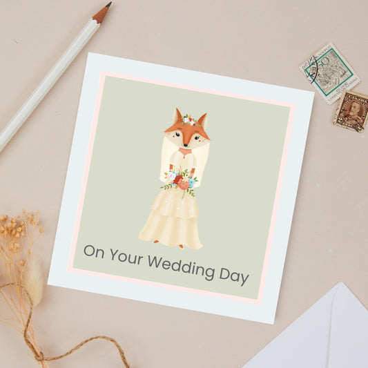 On your Wedding Day Card. This is a from me to you  Greeting card . The card features a Girl fox, dressed in a cream bridal dress. The background of the card is a light green. Set against a light beige background with stationery.
