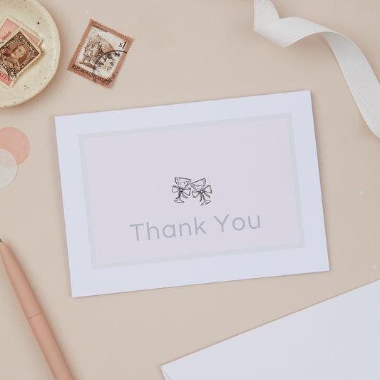 White card with 'Thank You' text and Champagne bow glasses design set against a light pink background with a grey border. The Card is set against a beige background, with a cream ribbon, and stationery.