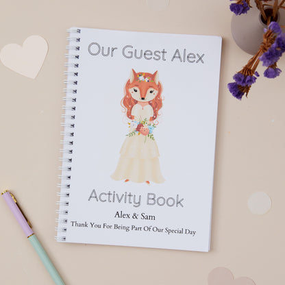 Children's Wedding activity book with an Auburn haired girl  fox dressed in bridal dress. She is holding a floral bouquet.
The illustration is on a beige background with stationery and purple dried flowers in the background